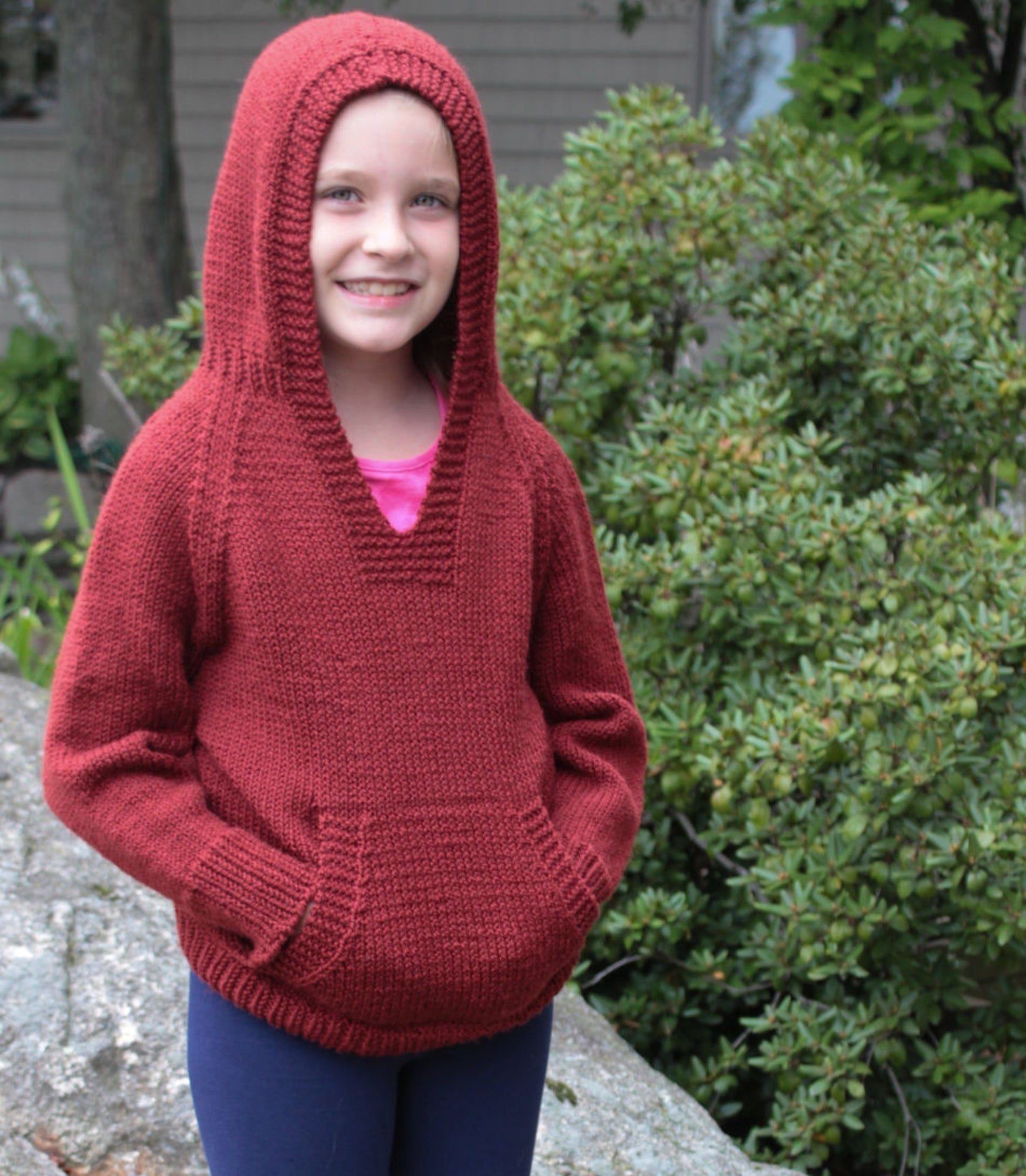 A smiling young girl stands outdoors, wearing a red Cottage Creations Wonderful Wallaby For Grown-Ups knitted hooded sweatshirt with her hands in the front hand-warming pockets. She has blue pants on and is standing in front of greenery and a rock.