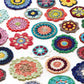 A collection of vibrant, intricately designed pieces from the “Crochet Kaleidoscope” by Ingram Content, laid out on a white background. The pieces feature a variety of patterns and yarn color combinations, including reds, blues, greens, and yellows, showcasing an array of circular and floral designs.