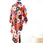 The Crochet Kaleidoscope by Ingram Content, a colorful crocheted shawl with hexagonal patterns in vibrant yarn colors of red, pink, orange, blue, and white, is draped over a mannequin. The shawl features intricate floral crocheted motifs on each hexagon and contrasts beautifully against the plain white background of the room.