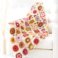 A white armchair is draped with Ingram Content's Crochet Kaleidoscope, a vibrant blanket featuring colorful hexagonal patterns. The blanket, showcasing various yarn shades like red, orange, yellow, green, pink, and purple, adds warmth to the light and airy room with its minimalist decor and tall piece of abstract art.