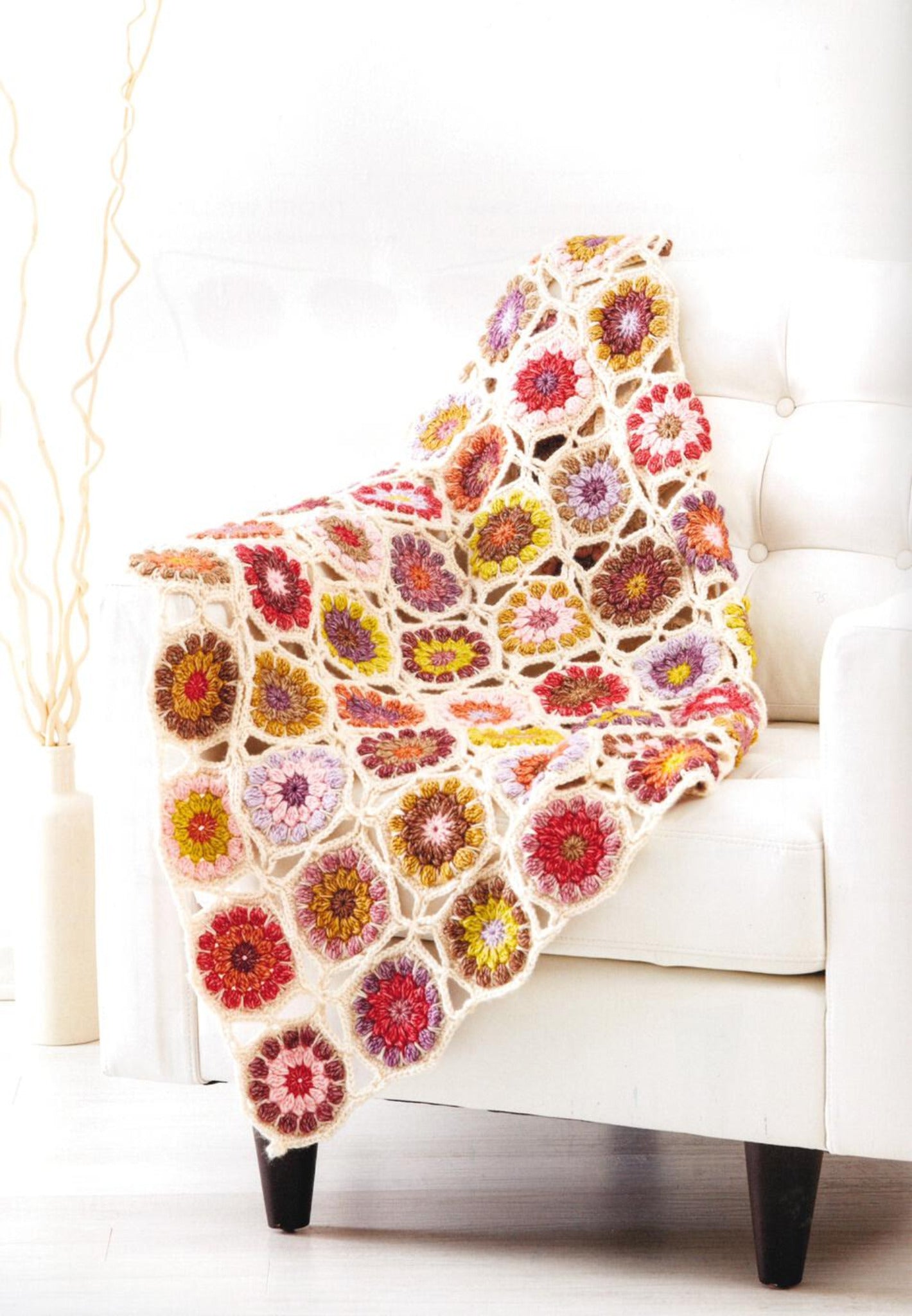 A white armchair is draped with Ingram Content's Crochet Kaleidoscope, a vibrant blanket featuring colorful hexagonal patterns. The blanket, showcasing various yarn shades like red, orange, yellow, green, pink, and purple, adds warmth to the light and airy room with its minimalist decor and tall piece of abstract art.