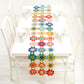 A white dining table set includes four white chairs, each outfitted with a white mug, bowl, and spoon. The center of the table is adorned with the Crochet Kaleidoscope by Ingram Content - a colorful, intricate crochet table runner that showcases vibrant crocheted motifs in various yarn colors. The setting is bright and minimalistic.