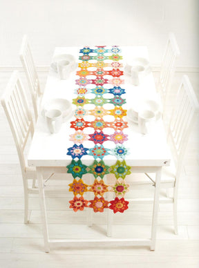 A white dining table set includes four white chairs, each outfitted with a white mug, bowl, and spoon. The center of the table is adorned with the Crochet Kaleidoscope by Ingram Content - a colorful, intricate crochet table runner that showcases vibrant crocheted motifs in various yarn colors. The setting is bright and minimalistic.