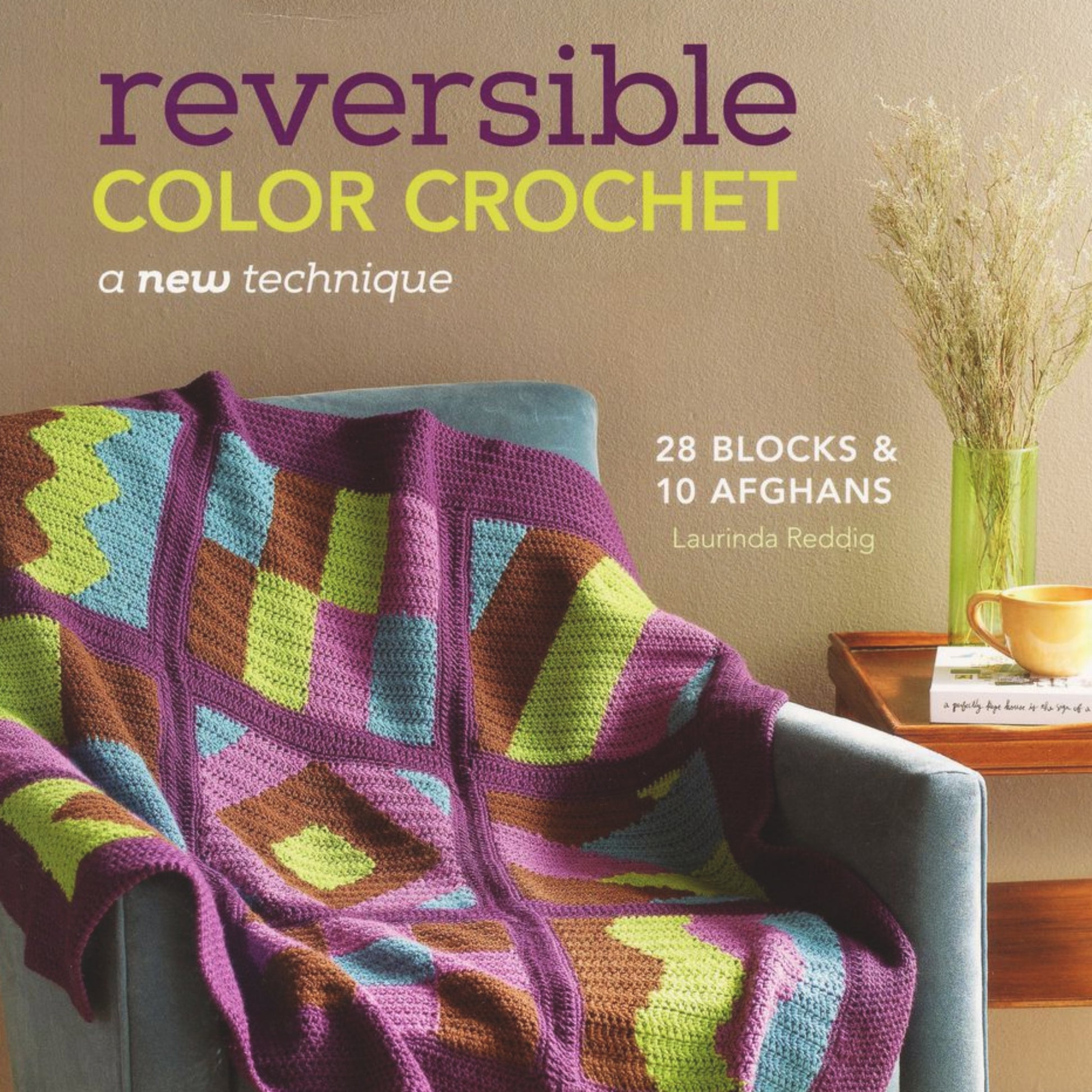 A crochet quilt adorned in vibrant, multicolored zigzag and geometric patterns is draped over a charcoal-colored chair. On the adjacent wooden table sits a green vase with dried flowers and an orange mug. The book cover reads "Reversible Color Crochet - A New Technique" by Ingram Content, perfect for afghan projects.