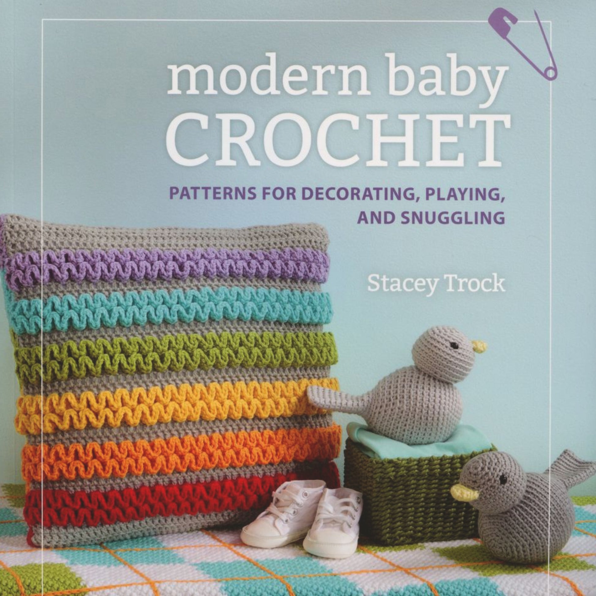 A book cover titled "Modern Baby Crochet - Patterns for Decorating, Playing, and Snuggling" by Stacey Trock and published by Martingale & Co features a stack of colorful crocheted pillows, two crocheted ducklings, and a pair of baby shoes on a multicolored blanket—perfect for your next crochet baby projects or baby shower gift ideas.