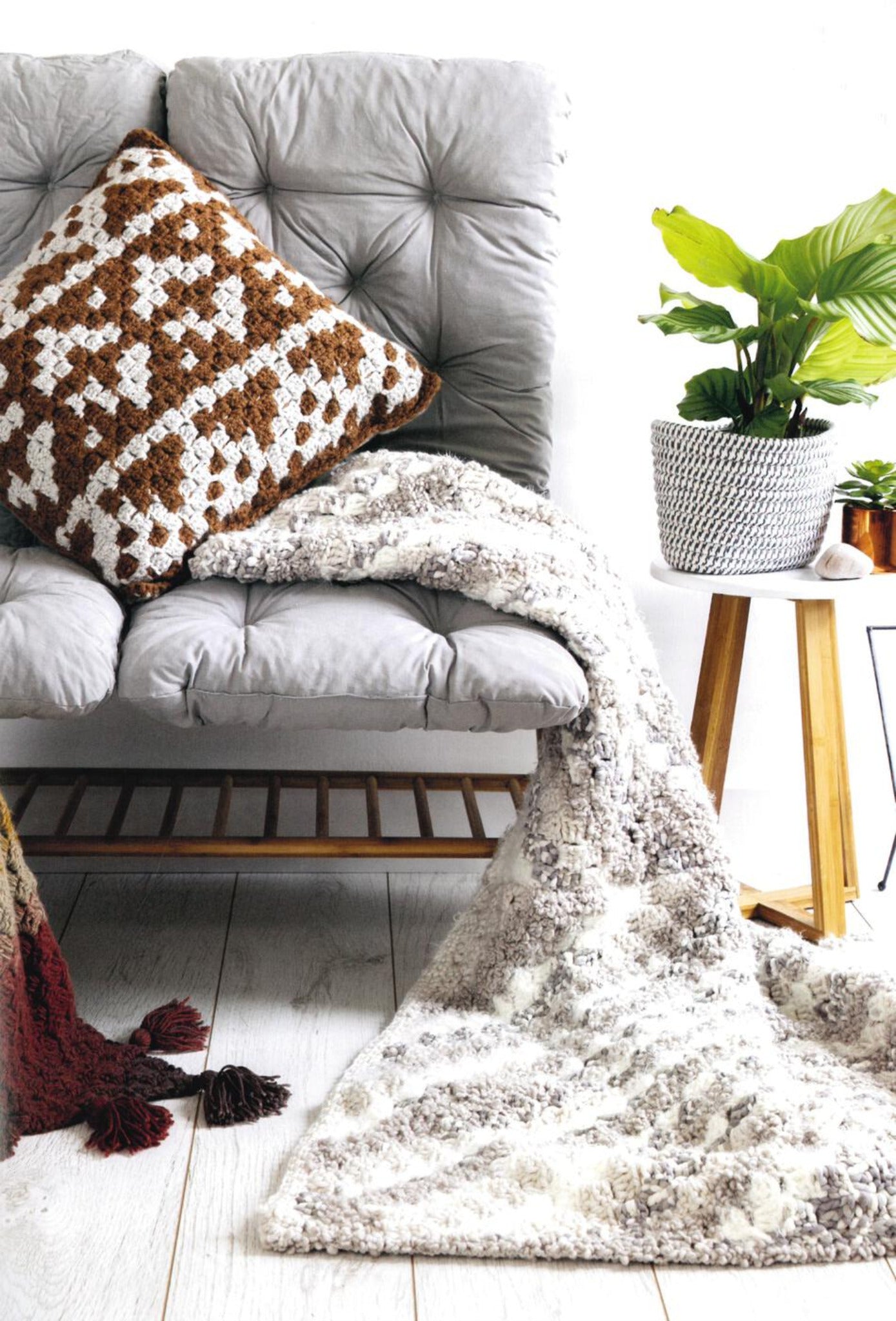 A cozy corner features a gray cushioned seat adorned with a brown and white geometric-patterned pillow. Draped over the seat is a stylish white and gray knitted blanket, created using techniques from "Corner to Corner Crochet - 15 Contemporary C2C Projects" by Ingram Content. Nearby, a small wooden table holds a potted green plant, adding a touch of nature to the home decor.