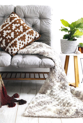 A cozy corner features a gray cushioned seat adorned with a brown and white geometric-patterned pillow. Draped over the seat is a stylish white and gray knitted blanket, created using techniques from "Corner to Corner Crochet - 15 Contemporary C2C Projects" by Ingram Content. Nearby, a small wooden table holds a potted green plant, adding a touch of nature to the home decor.