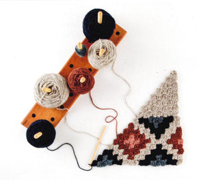 The "Corner to Corner Crochet - 15 Contemporary C2C Projects" by Ingram Content features an elegant wooden holder with five spools of yarn in blue, grey, cream, red, and black. A crochet hook is attached to a partially completed C2C crochet granny square triangle blanket in shades of grey, red, black, and cream—perfect for adding a touch of charm to your home decor.