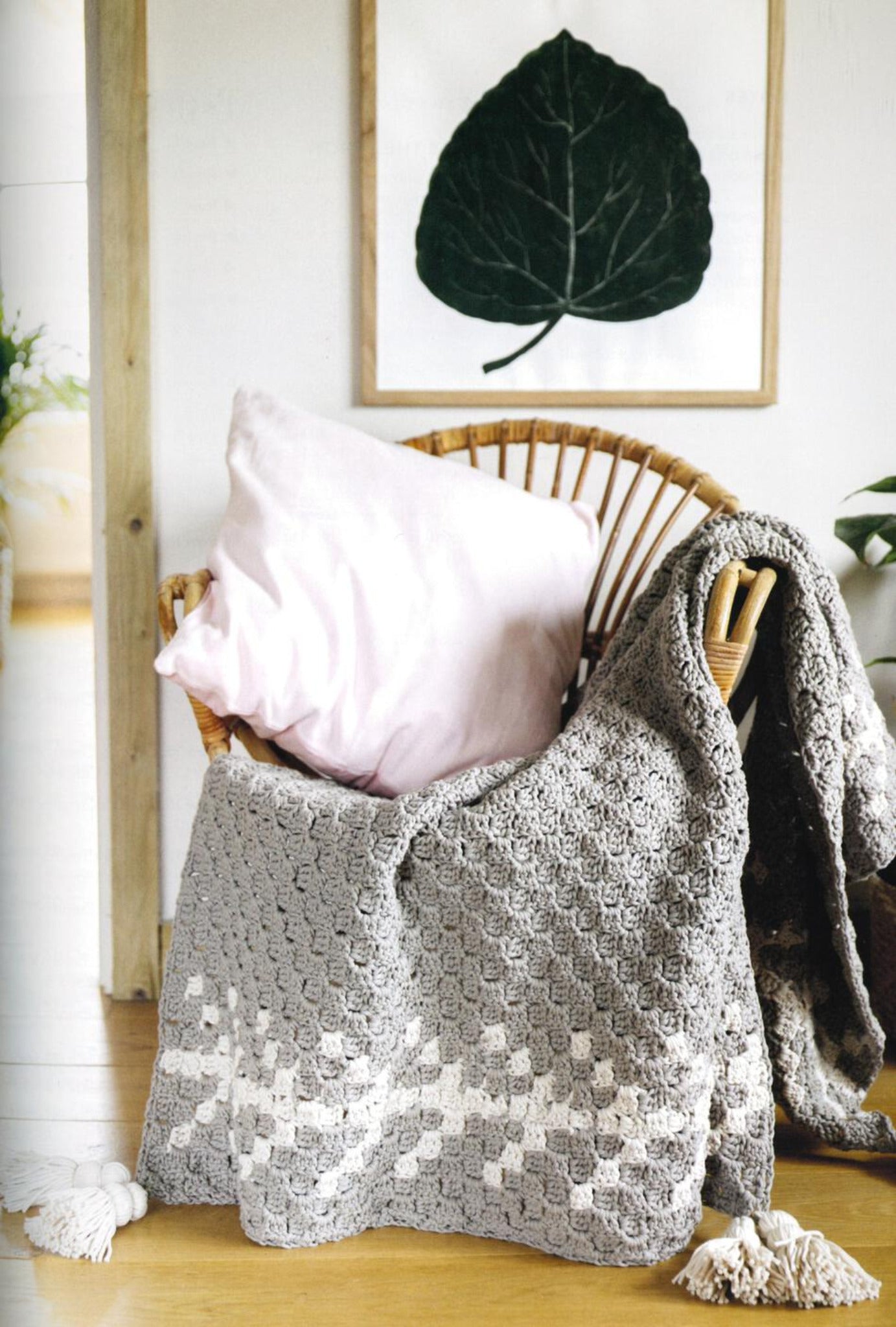 A cozy corner featuring a wicker chair draped with the Corner to Corner Crochet blanket from Ingram Content, showcasing a beautiful gray and white C2C design, along with a light pink pillow, is shown. A framed leaf artwork hangs on the wall behind the chair, and a green plant is visible in the background on the left, adding to the charming home decor.