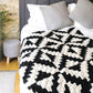 A cozy bedroom features a bed adorned with a blanket inspired by the "Corner to Corner Crochet - 15 Contemporary C2C Projects" from Ingram Content, two gray pillows against a wooden accent wall, and a small round gray knitted stool. A potted plant and a light-colored rug complete the room's home decor.