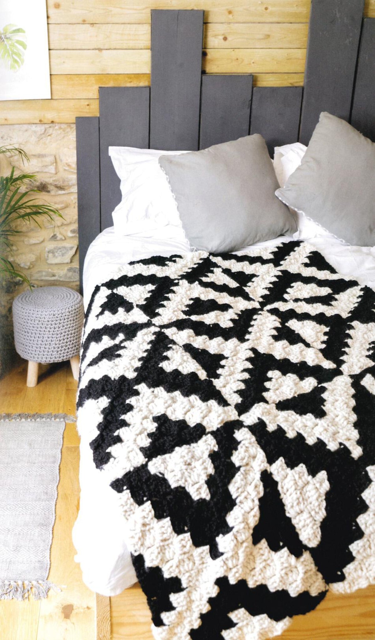 A cozy bedroom features a bed adorned with a blanket inspired by the "Corner to Corner Crochet - 15 Contemporary C2C Projects" from Ingram Content, two gray pillows against a wooden accent wall, and a small round gray knitted stool. A potted plant and a light-colored rug complete the room's home decor.