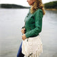 A woman with long blonde hair stands by a lake, wearing a green jacket and blue jeans. She holds the Carefree Crochet bag from Ingram Content, a stylish white wearable crochet bag adorned with fringe. The background reveals calm water and a distant wooded shoreline beneath a cloudy sky.