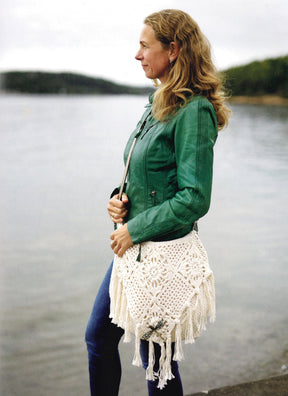 A woman with long blonde hair stands by a lake, wearing a green jacket and blue jeans. She holds the Carefree Crochet bag from Ingram Content, a stylish white wearable crochet bag adorned with fringe. The background reveals calm water and a distant wooded shoreline beneath a cloudy sky.