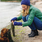 A woman wearing a green jacket, blue hat, and matching blue gloves crouches by the lakeshore, beaming at a small brown dog holding strands of seaweed in its mouth. The backdrop of a sandy beach and blurred treeline creates the perfect scene for enthusiasts of Carefree Crochet by Ingram Content.