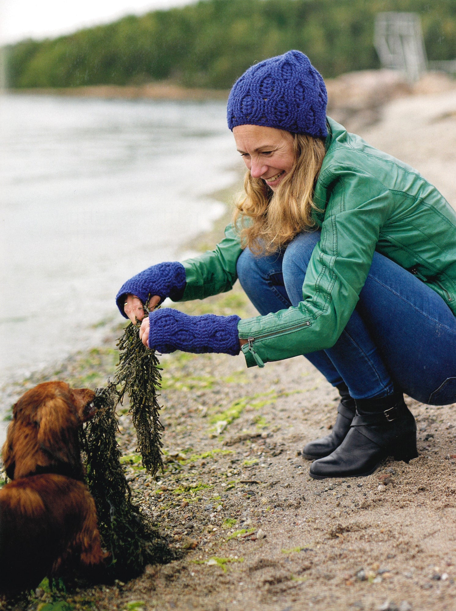 A woman wearing a green jacket, blue hat, and matching blue gloves crouches by the lakeshore, beaming at a small brown dog holding strands of seaweed in its mouth. The backdrop of a sandy beach and blurred treeline creates the perfect scene for enthusiasts of Carefree Crochet by Ingram Content.