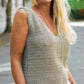 A woman with long, blonde hair is smiling outdoors. She is wearing the Carefree Crochet top by Ingram Content, a gray knitted sleeveless piece featuring dynamic openwork, along with blue jeans and several bracelets on her left wrist. She also has a long necklace and is standing on a paved surface with greenery in the background.