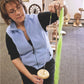 A person wearing glasses and a light blue vest is demonstrating wool spinning using Halcyon Yarn's Drop Spindle How-To. They hold a strand of green wool and are carefully twisting it into yarn, with various Halcyon Yarn spinning kits visible in the background.