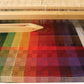 A wooden 8-shaft loom holds the "Color Gamp - A Study in Color & Weave, 10/2 Cotton" by Halcyon Yarn, featuring intricately woven multicolored fabric that transitions from green to blue, red, and purple alongside black-and-white patterns. Resting on the loom is a wooden shuttle, emphasizing the textile creation process using 10/2 Pearl Cotton.