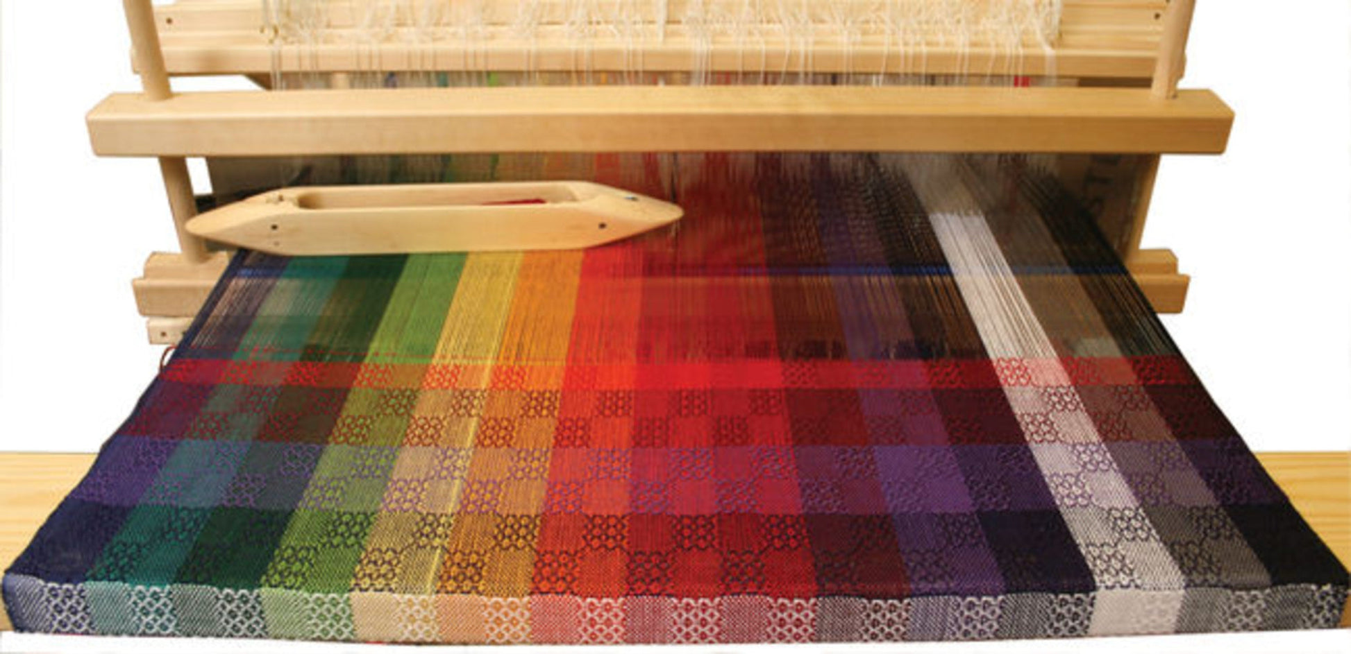 A wooden 8-shaft loom holds the "Color Gamp - A Study in Color & Weave, 10/2 Cotton" by Halcyon Yarn, featuring intricately woven multicolored fabric that transitions from green to blue, red, and purple alongside black-and-white patterns. Resting on the loom is a wooden shuttle, emphasizing the textile creation process using 10/2 Pearl Cotton.