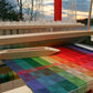 A Halcyon Yarn Color Gamp - A Study in Color & Weave, 10/2 Cotton wooden 8-shaft loom is pictured outdoors at sunset, weaving a vibrant, gradient-patterned fabric. The fabric transitions smoothly through various colors, creating a visually striking effect against the backdrop of bare trees and a colorful sky.