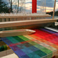 A vibrantly colored textile in a rainbow gradient of green, red, blue, and yellow hues is being woven on a loom kit. A wooden shuttle sits elegantly on the setup. In the background, a picturesque sunset over a lake with silhouetted trees creates an ideal setting for any weaving study using Halcyon Yarn's Color Gamp - A Study in Color & Weave, 10/2 Cotton digital product.