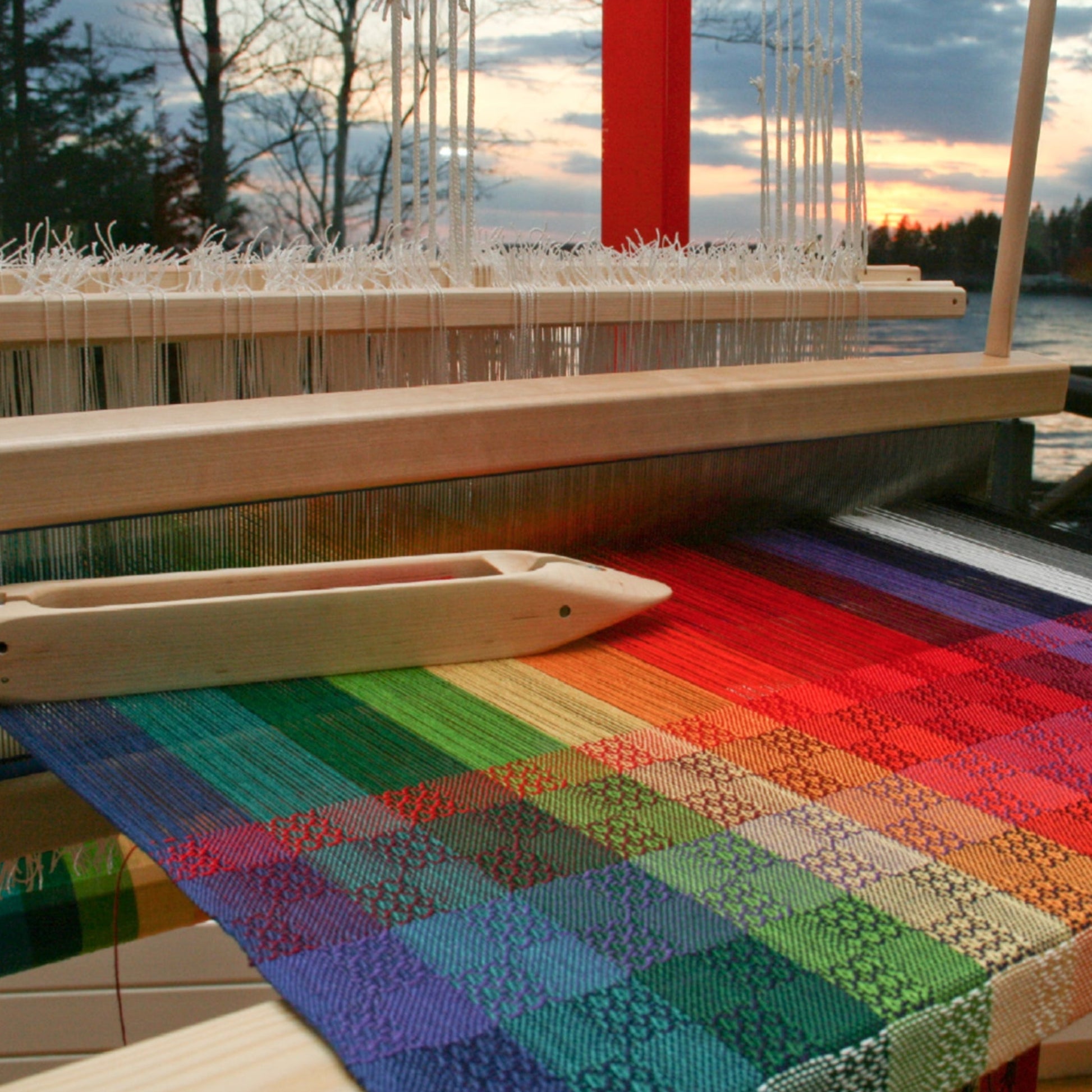 A vibrantly colored textile in a rainbow gradient of green, red, blue, and yellow hues is being woven on a loom kit. A wooden shuttle sits elegantly on the setup. In the background, a picturesque sunset over a lake with silhouetted trees creates an ideal setting for any weaving study using Halcyon Yarn's Color Gamp - A Study in Color & Weave, 10/2 Cotton digital product.