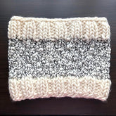 Presenting the Aguas Blancas Cowl Pattern by Kristel Nieves, a snug knitted creation made from exquisite Merino wool. It features wide cream-colored bands at both the top and bottom, complemented by a central section of black and white speckled yarn. The intricate design is chunky and elegantly raised.