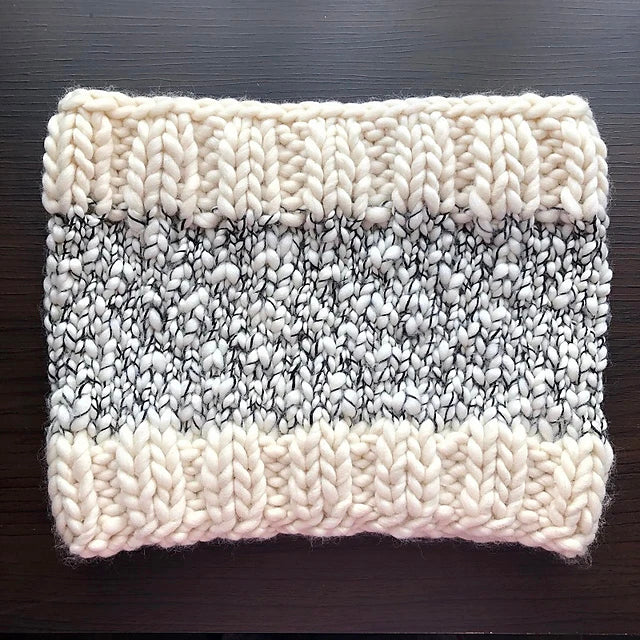 Presenting the Aguas Blancas Cowl Pattern by Kristel Nieves, a snug knitted creation made from exquisite Merino wool. It features wide cream-colored bands at both the top and bottom, complemented by a central section of black and white speckled yarn. The intricate design is chunky and elegantly raised.