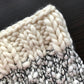 Close-up of a knitted fabric featuring a thick, cream-colored yarn intertwined with black strands, reminiscent of the Aguas Blancas Cowl Pattern by Kristel Nieves. The texture is chunky with prominent stitches, crafted from Malabrigo merino wool, resting on a dark, textured surface.