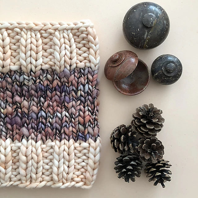 An Aguas Blancas Cowl Pattern by Kristel Nieves, crafted from merino wool in beige, brown, and purple hues, is displayed next to decorative round stones, a small metallic container, and a collection of pine cones on a light surface.