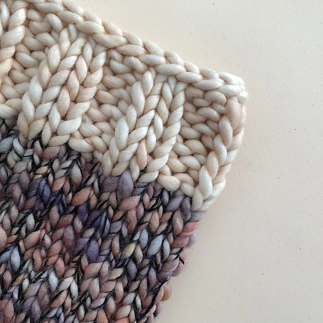 A detailed view of a knitted fabric showcasing a chunky yarn style made from Malabrigo merino wool. The knit shifts from a light beige at the top to an intricate blend of beige, brown, and purple tones, emphasizing its texture and pattern against a light backdrop, reminiscent of the Aguas Blancas Cowl Pattern by Kristel Nieves.