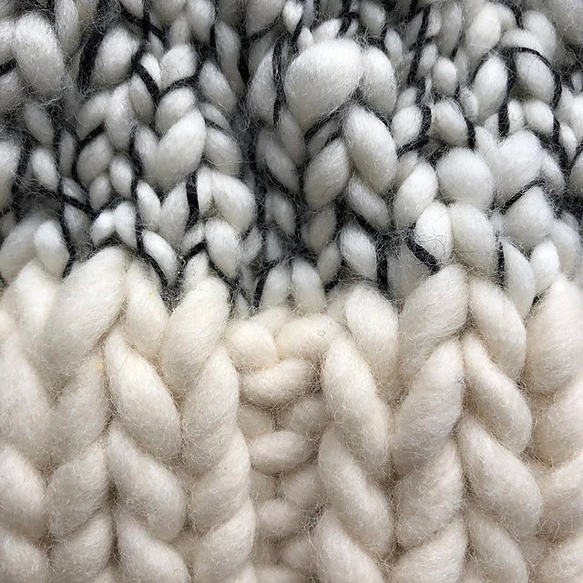 A close-up of chunky knitted fabric in a blend of white, cream, and gray yarns highlights the rich texture of merino wools. The thick stitches create a cozy appearance with visible strands of darker yarn woven throughout, reminiscent of the Aguas Blancas Hat Pattern by Kristel Nieves.