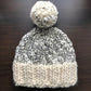 The Aguas Blancas Hat Pattern by Kristel Nieves is a cozy knitted beanie crafted from soft merino wool, complete with a large pom-pom on top. It features an intricate black and white speckled design and a solid white brim, beautifully portrayed against a dark wooden background.