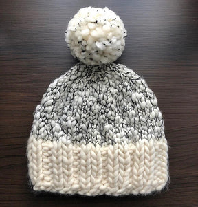 The Aguas Blancas Hat Pattern by Kristel Nieves is a cozy knitted beanie crafted from soft merino wool, complete with a large pom-pom on top. It features an intricate black and white speckled design and a solid white brim, beautifully portrayed against a dark wooden background.