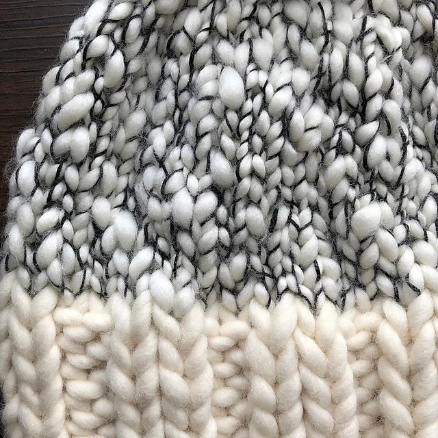 Close-up of a knitted fabric featuring a blend of white and black chunky yarn in the upper section and solid cream yarn in the lower, crafted with luxurious merino wools. The thick texture evokes the cozy charm of an Aguas Blancas Hat Pattern by Kristel Nieves.