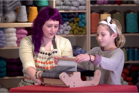 A woman with purple hair and a girl with a headband are working together at a weaving loom in a yarn store. Shelves filled with colorful yarn balls are in the background. The woman is demonstrating the process while the girl listens attentively.