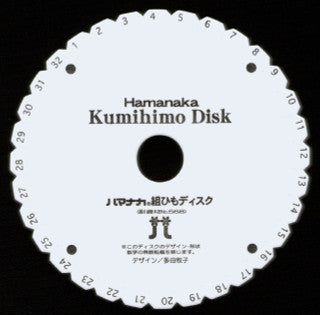 A round white Kumihimo disk labeled "BraidersHand Kumihimo Disk" features evenly spaced numbered slots from 1 to 32 around the outer edge. Japanese text and two icons of braiding tools are printed in the center. Perfect for creating braids, this portable project tool, marketed under the Braiders Hand/Saunders brand, is essential for crafting kumihimo cords.