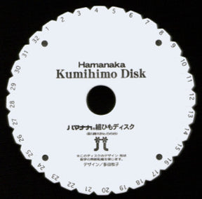 A round white Kumihimo disk labeled "BraidersHand Kumihimo Disk" features evenly spaced numbered slots from 1 to 32 around the outer edge. Japanese text and two icons of braiding tools are printed in the center. Perfect for creating braids, this portable project tool, marketed under the Braiders Hand/Saunders brand, is essential for crafting kumihimo cords.