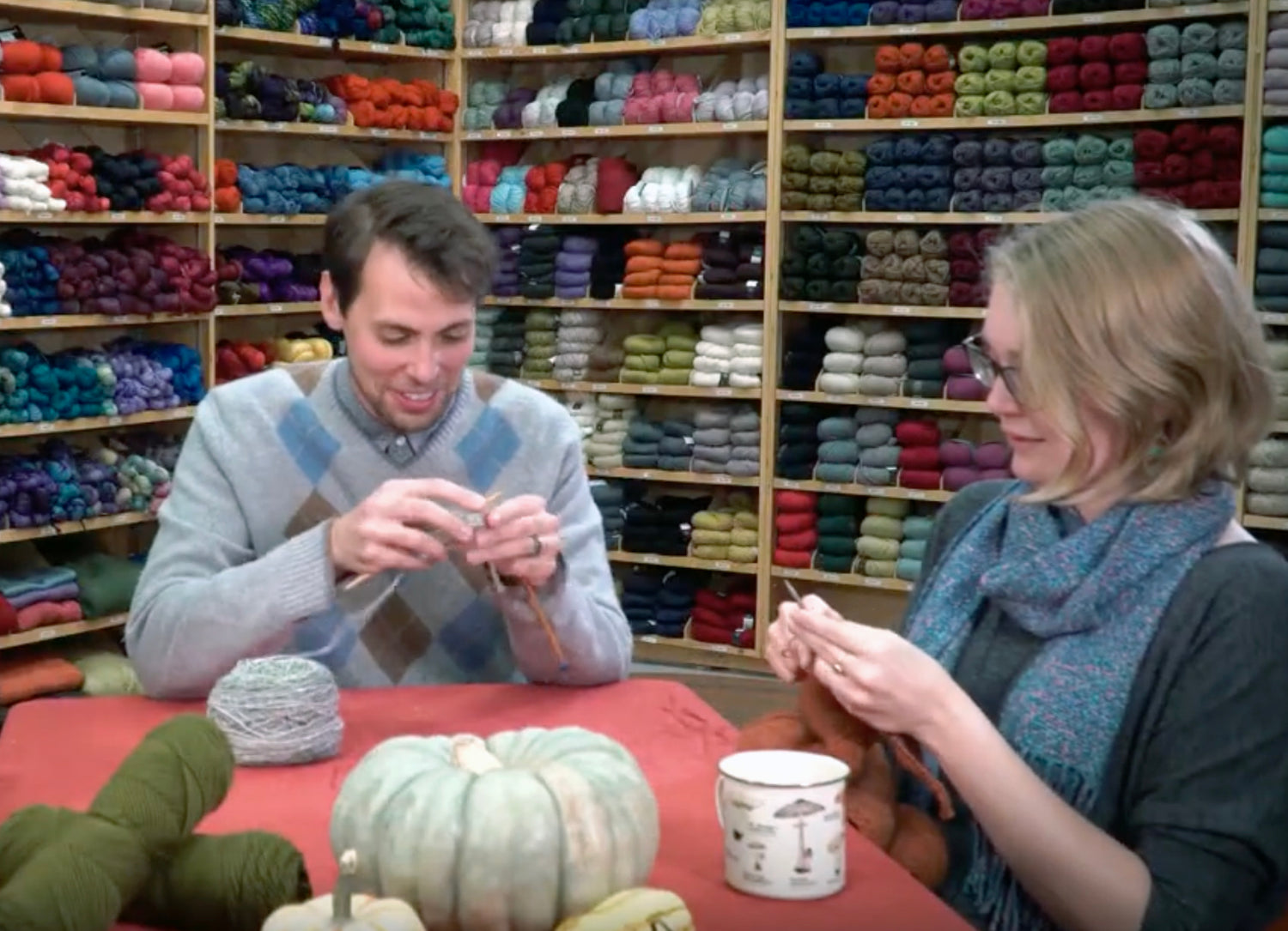 Two people sit at a table in a yarn store, knitting with various colored yarns. Shelves filled with neatly stacked yarns are in the background. A pumpkin and a mug are on the table, adding a cozy touch to the scene.