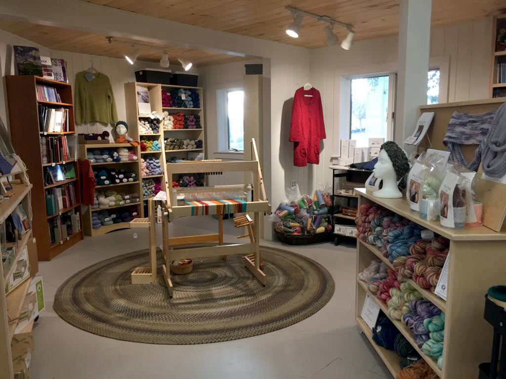 A cozy yarn shop features shelves filled with colorful yarns and knitting supplies. In the center of the room is a wooden weaving loom on a braided round rug. Finished garments and knitting books line the walls, illuminated by warm overhead lighting.