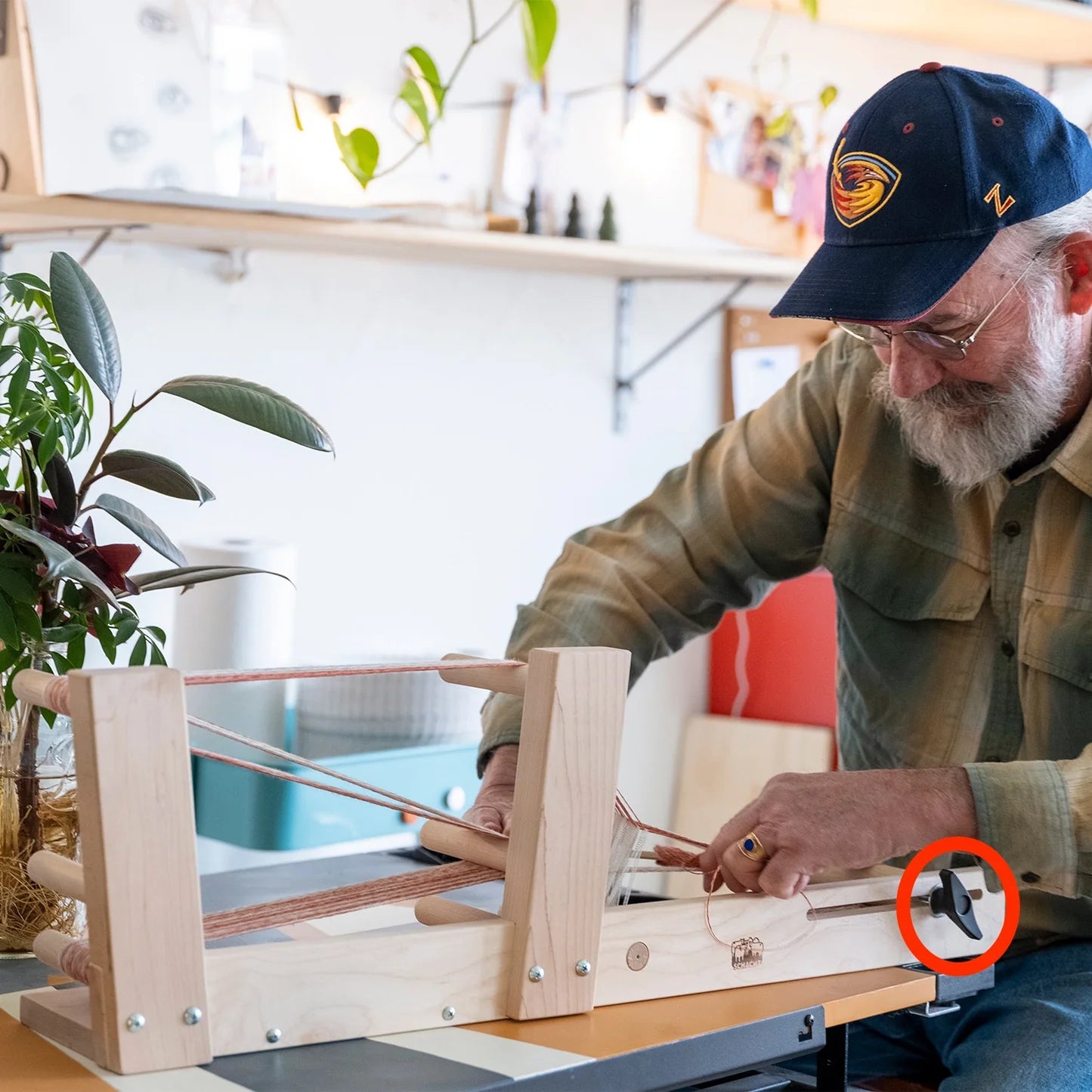 A person wearing a plaid shirt and a cap is using an Inkle Loom T-Knob by Schacht Spindle Co. to weave. They are seated at a table, focused on their work. There is a green plant to the left and shelves with various items in the background. A red circle highlights a black part of the loom.