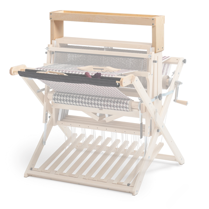 The Schacht High Castle Tray by Schacht Spindle Co. is a wooden handloom weaving machine made from light-colored wood, equipped with various levers and rollers. It prominently displays intricately woven fabric and features detailed mechanical components essential for weaving textiles, including a built-in tray for the convenient storage of tools.