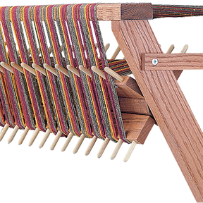 A close-up view of a wooden weaving loom with multicolored threads. The Schacht Sectional Warp Beam for Standard Floor Loom, by Schacht Spindle Co., features evenly spaced pegs, angled supports, and a sectional warp beam, showcasing a traditional crafting tool used for weaving textiles.