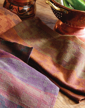 A close-up of colorful woven fabric, from the Best of Handwoven Yarn Series: A Dozen Projects in 8/2 Cotton by Long Thread Media, and a copper colander containing green beans. The woven fabrics, possibly patterned napkins, display various earth-toned patterns in shades of orange, brown, and purple. The setting appears to be a wooden table with additional copper kitchenware.