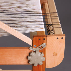 Close-up of an Ashford Second Back Beam in use on a table loom. The loom has multiple white and a few black threads stretched tightly across its frame. A beige adjustment knob with the word "Ashford" is visible on the left side, and an Ashford Second Back Beam from Ashford Handicrafts Limited helps maintain tension on the threads.