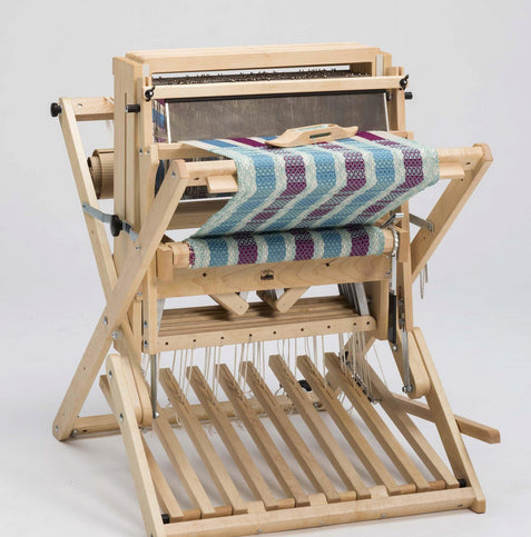 A wooden Schacht 18" Wolf Pup loom from Schacht Spindle Co. stands against a plain background, showcasing a partially woven textile with a zigzag pattern in blue, turquoise, and burgundy. The loom's structure is made of light-colored wood, featuring various levers, beams, and threads visible along with the Schacht Heddle bars.