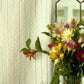 A bouquet of colorful flowers, featuring daisies and lilies, is elegantly arranged in a glass vase on a windowsill. The window's patterned glass pane complements the light-colored curtains adorned with thin vertical stripes. Shafts of natural light highlight a printed version of Long Thread Media's "Best of Handwoven Yarn Series: A Dozen Projects in 10/2 Pearl Cotton" that sits nearby, casting a serene ambiance over the scene.