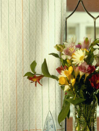 A bouquet of colorful flowers, featuring daisies and lilies, is elegantly arranged in a glass vase on a windowsill. The window's patterned glass pane complements the light-colored curtains adorned with thin vertical stripes. Shafts of natural light highlight a printed version of Long Thread Media's "Best of Handwoven Yarn Series: A Dozen Projects in 10/2 Pearl Cotton" that sits nearby, casting a serene ambiance over the scene.