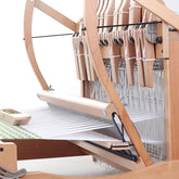 A Shuttle Race Add-On Kit from Ashford Handicrafts Limited's Ashford Table Looms collection features white threads stretched across the loom. A shuttle swiftly moves along the green and white fabric being woven, while multiple heddles and a beater demonstrate the loom's detailed craftsmanship.