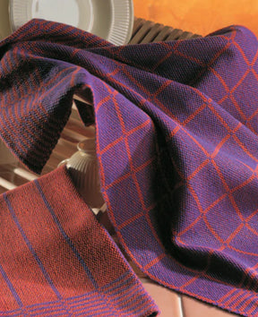 A close-up of two woven fabrics draped over dishes. The top fabric resembles a stylish tablecloth in purple with a red grid pattern, while the bottom one is red with dark blue diagonal stripes. The background showcases neutral-colored dishes in a warm, orange-tinted setting. These beautiful textiles are featured in the "Best of Handwoven Yarn Series: A Dozen Projects in 10/2 Pearl Cotton (Printed Version)" by Long Thread Media.